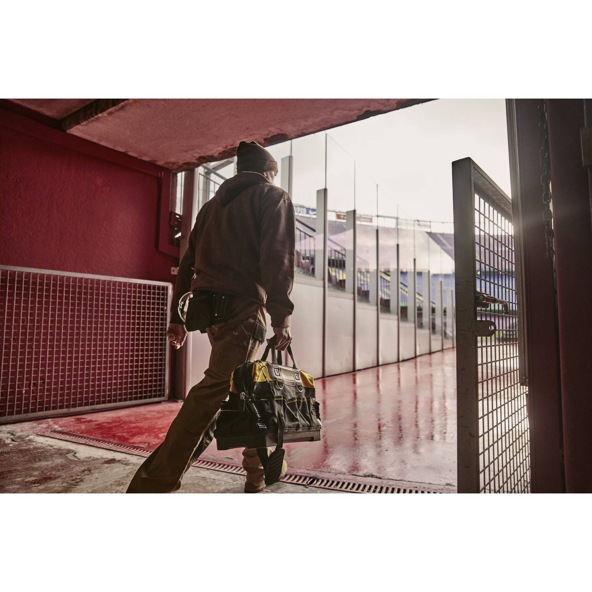 Eine Person in Arbeitskleidung trägt eine große Tasche durch ein Tor in einem Stadion. Der Boden ist rot, und es ist bewölkt.