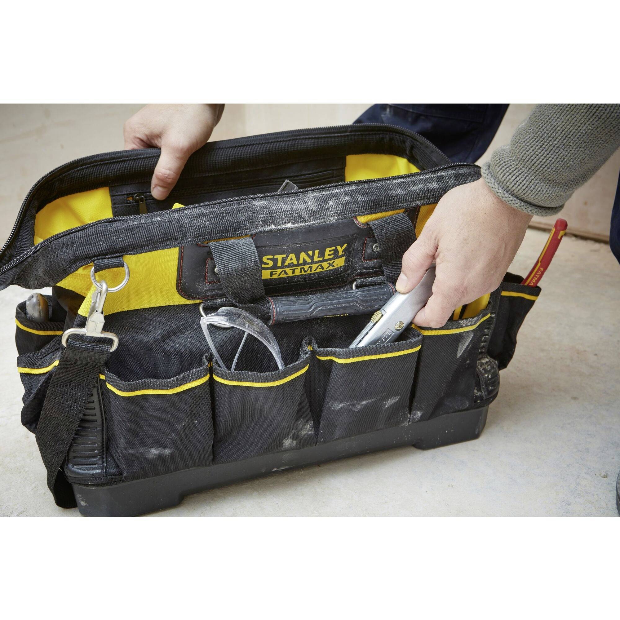 Eine Hand greift in eine geöffnete schwarze Werkzeugtasche mit gelben Akzenten und mehreren Fächern. Die Tasche enthält verschiedene Werkzeuge.