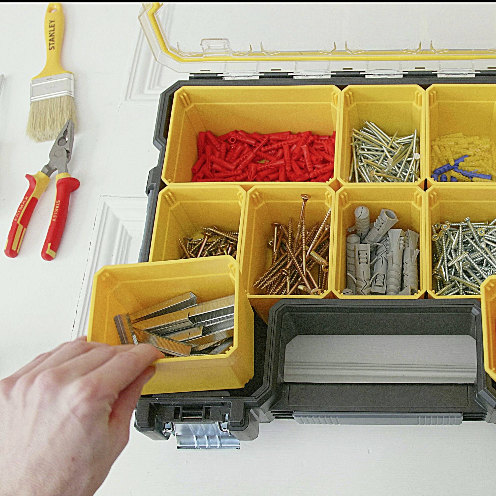 Ein geöffneter Werkzeugkasten enthält verschiedene Schrauben und Dübel, links daneben liegen Pinsel und Werkzeuge auf einem Tisch.