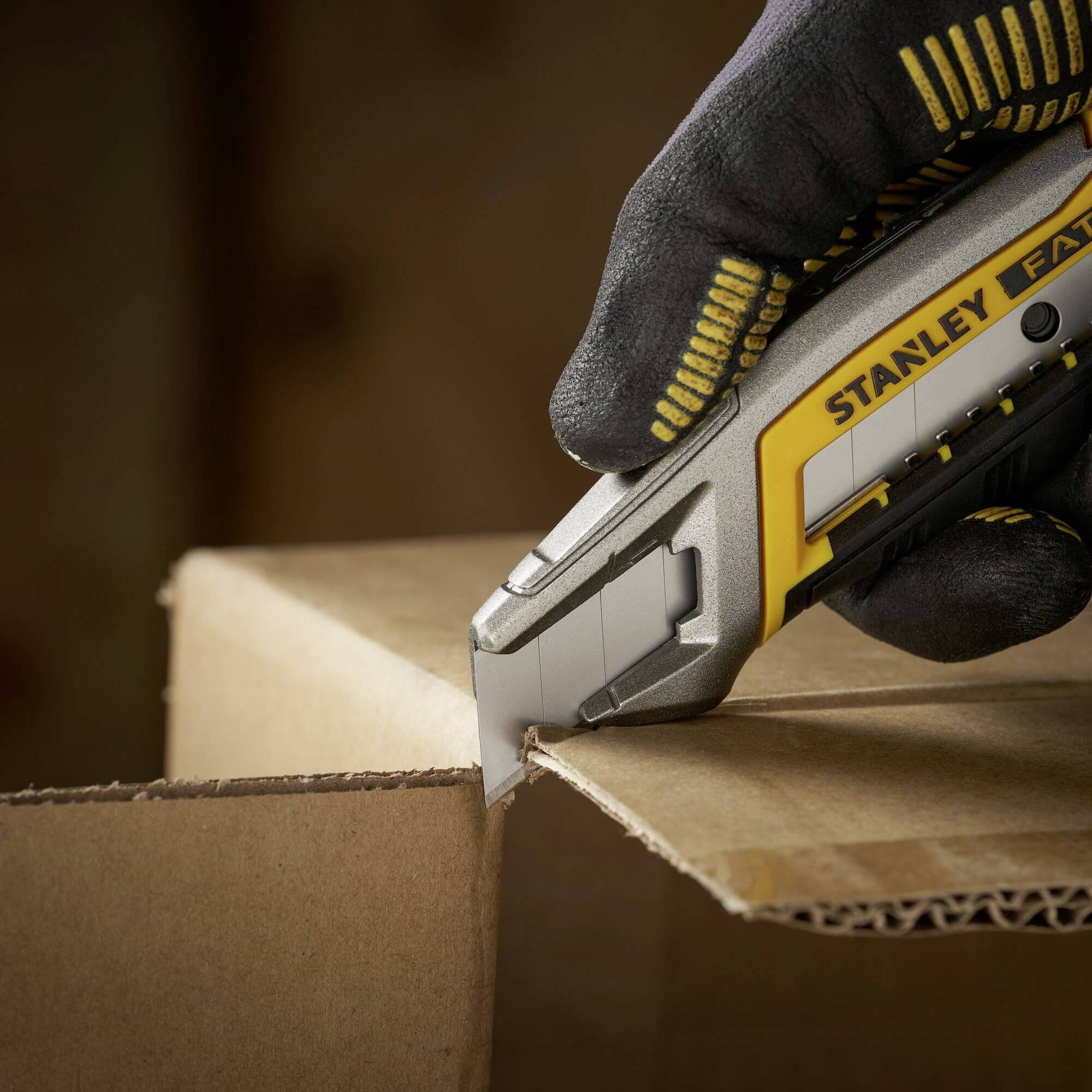 Eine behandschuhte Hand schneidet mit einem Stanley-Messer einen Karton an der Ecke auf.
