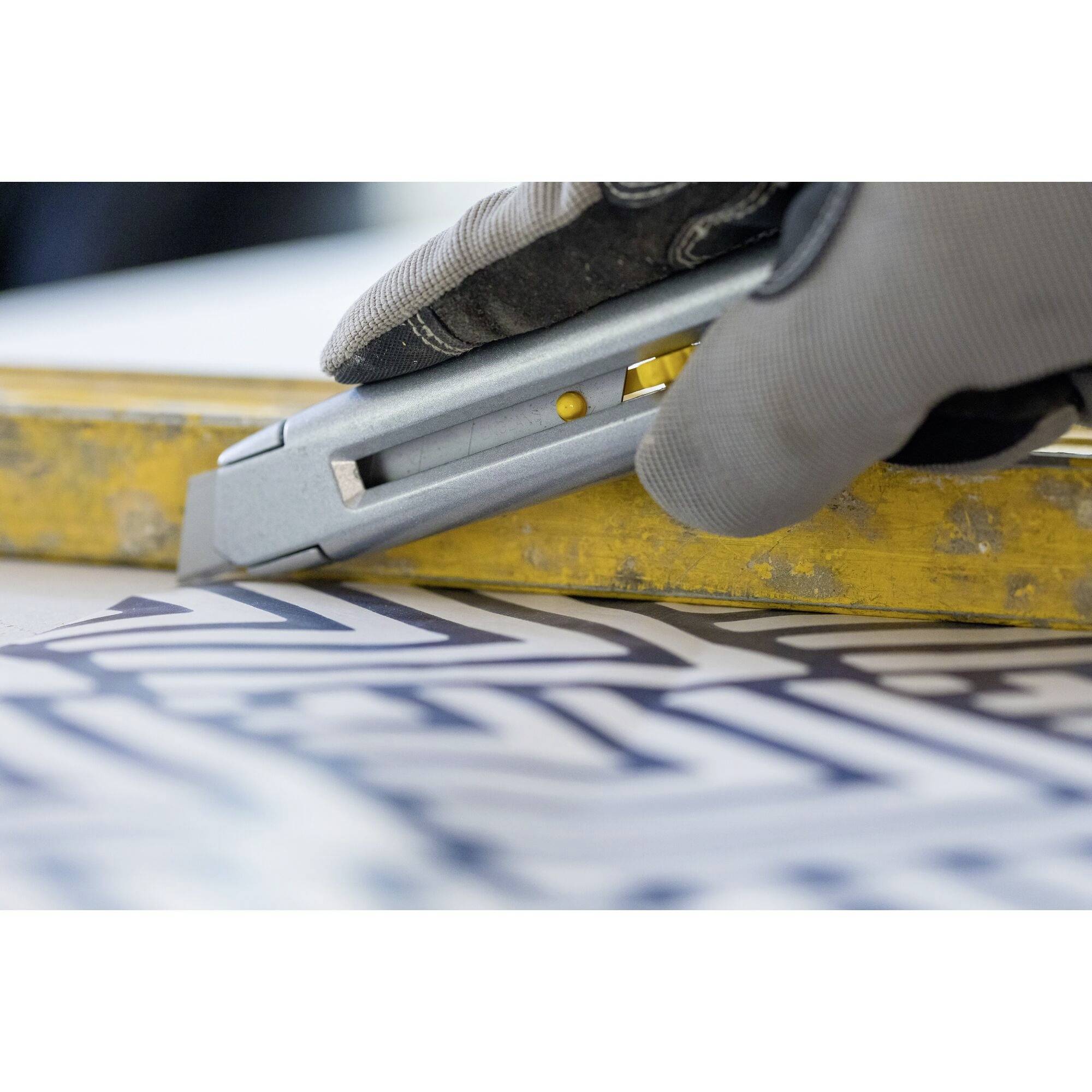 Hand mit Handschuh benutzt ein Cuttermesser, um ein Stück Papier mit geometrischem Muster auf einem gelben Lineal zuzuschneiden.