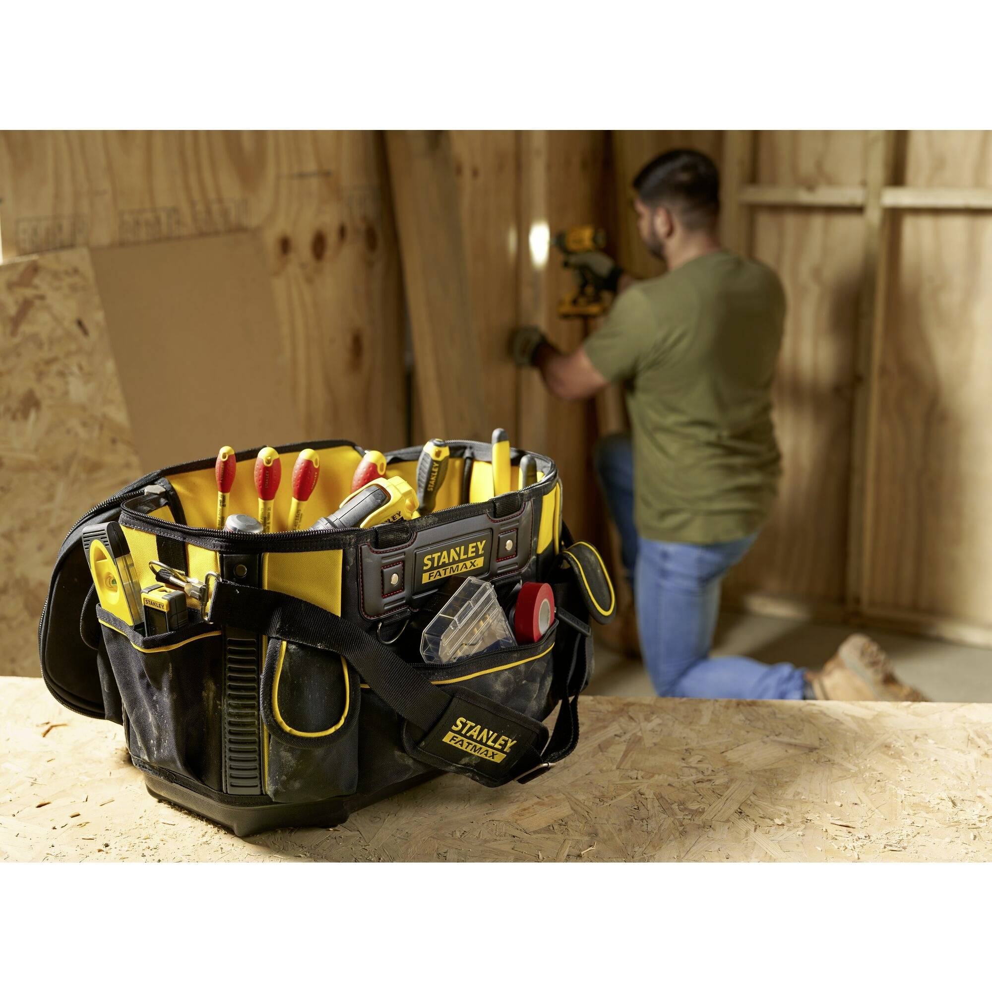Werkzeugtasche mit diversen Handwerkzeugen im Vordergrund. Unscharfe Person im Hintergrund arbeitet an einem Holzprojekt.