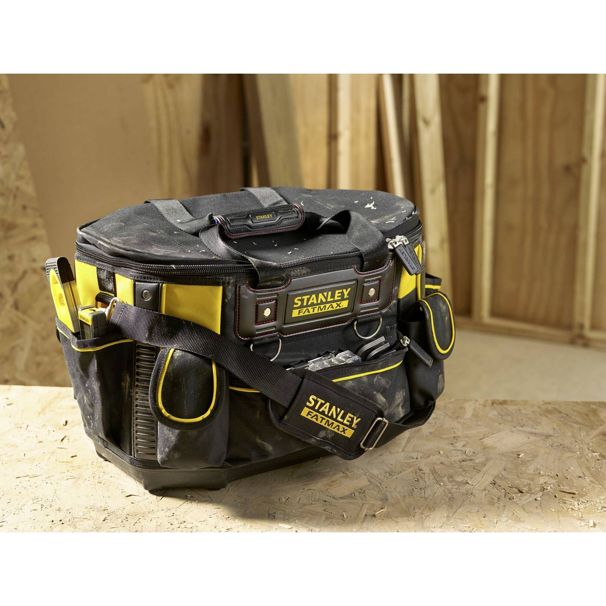 Eine schwarze Werkzeugtasche mit gelben Akzenten und mehreren Fächern steht auf einem Holztisch; der Hintergrund zeigt Holzwände.