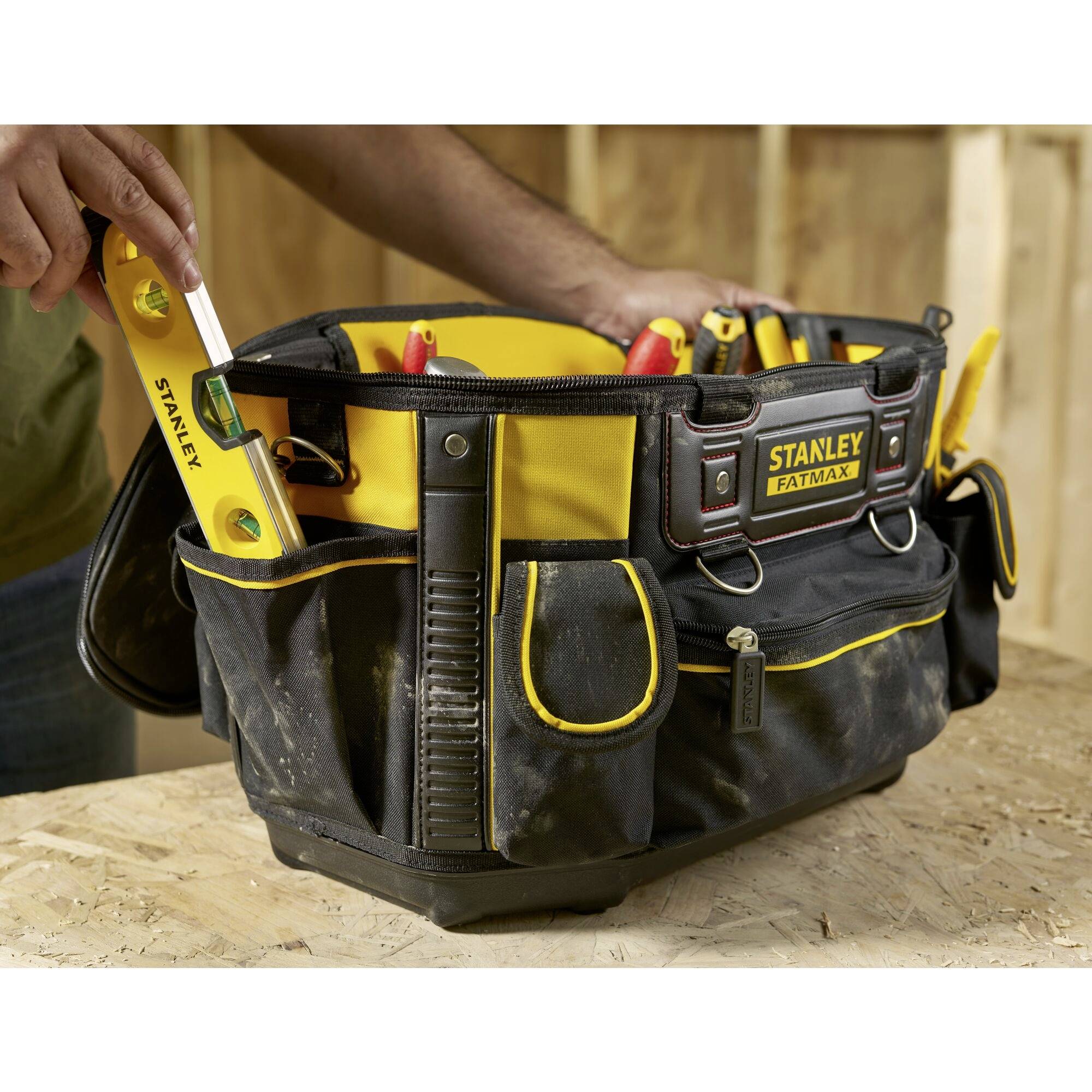 Eine Person legt eine gelbe Wasserwaage in eine schwarze Werkzeugtasche auf einer Holzoberfläche. Die Tasche ist voll mit Werkzeugen.