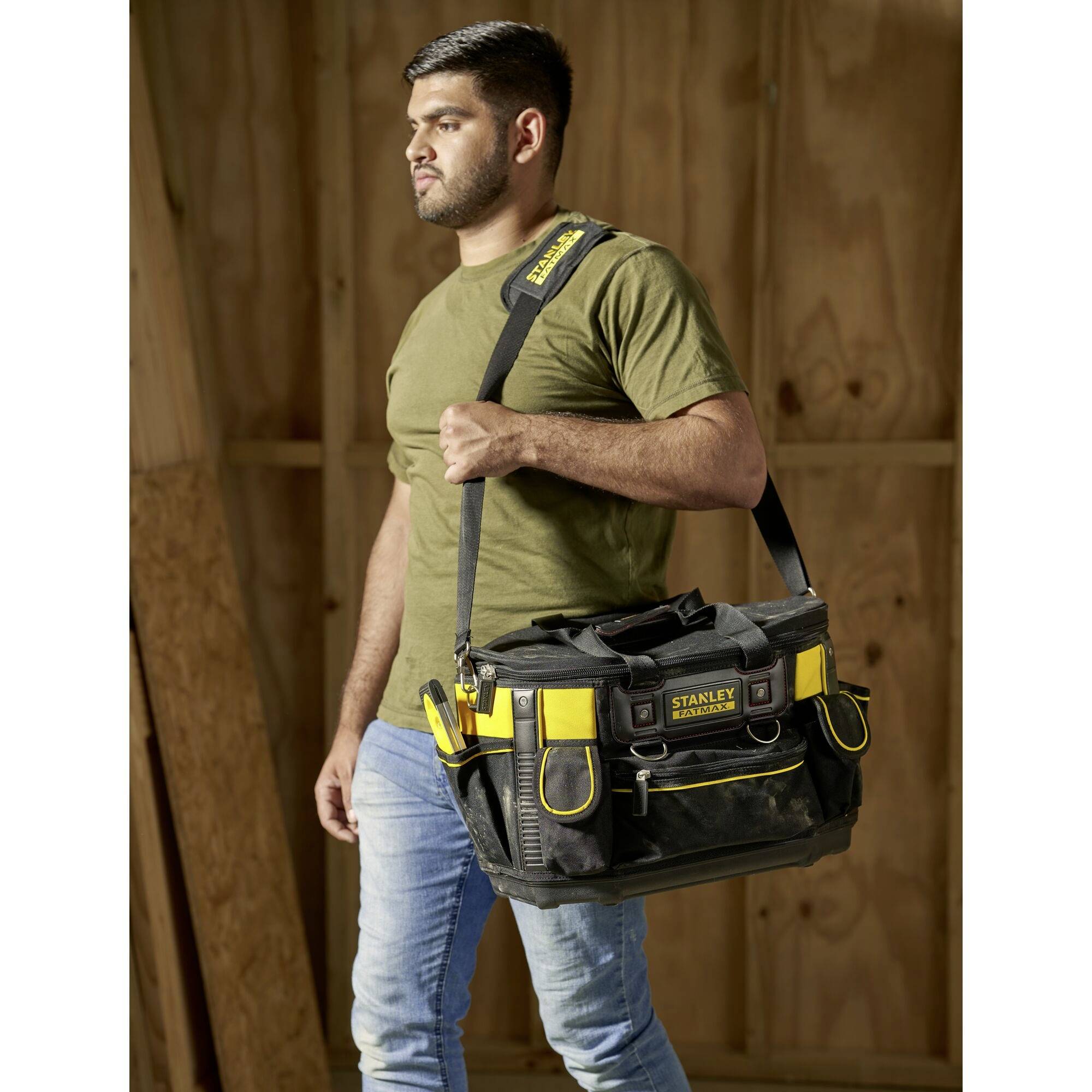 Ein Mann trägt eine schwarze Werkzeugtasche mit gelben Akzenten. Die Tasche hat mehrere Fächer und der Mann trägt ein olivgrünes T-Shirt.