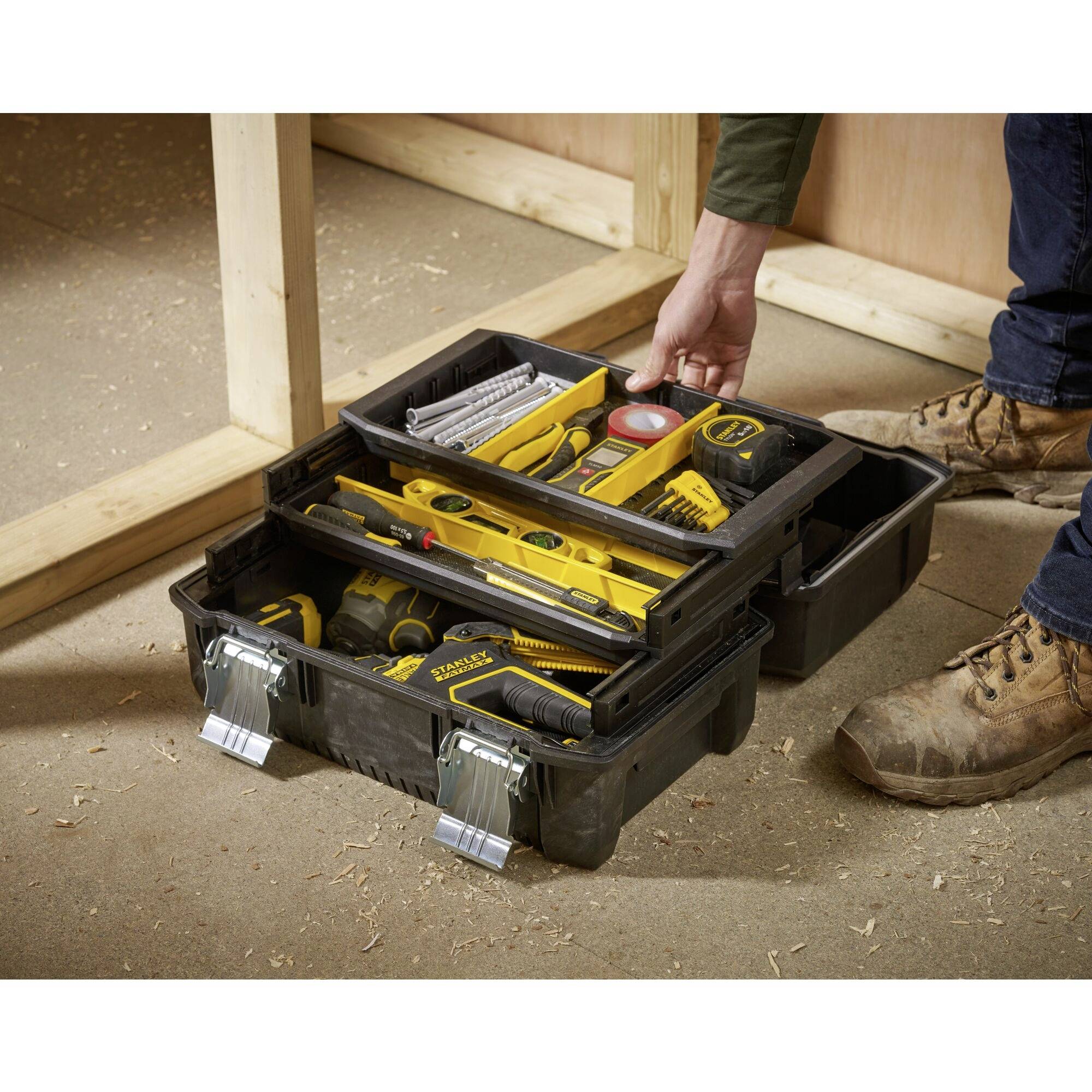 Ein geöffneter Werkzeugkasten auf dem Boden mit gelben Handwerkzeugen und Zubehör. Eine Person in Arbeitskleidung greift nach Werkzeugen.