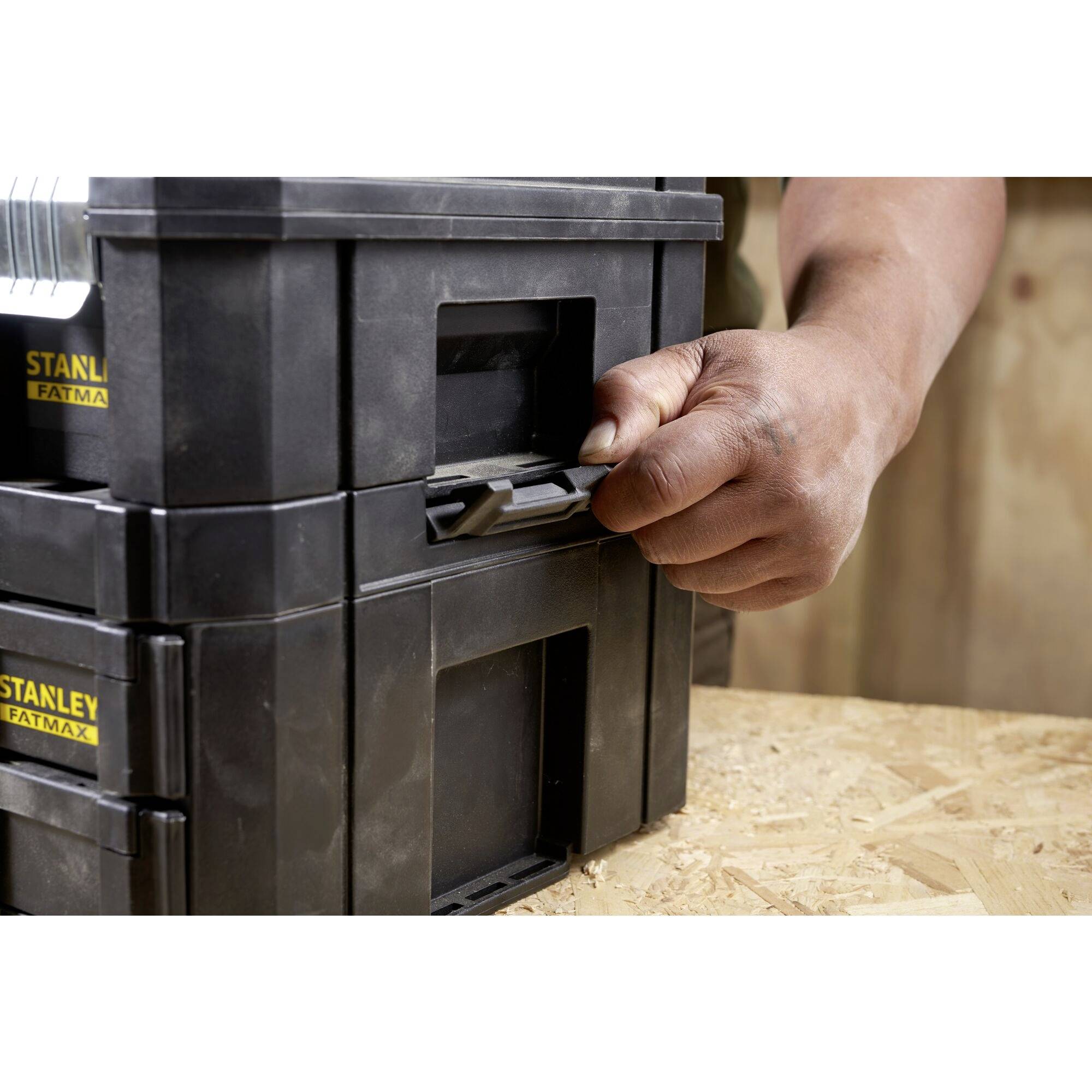Eine Person öffnet mit der Hand eine schwarze, stapelbare Werkzeugbox der Marke Stanley Fatmax. Sie steht auf einer Holzoberfläche.