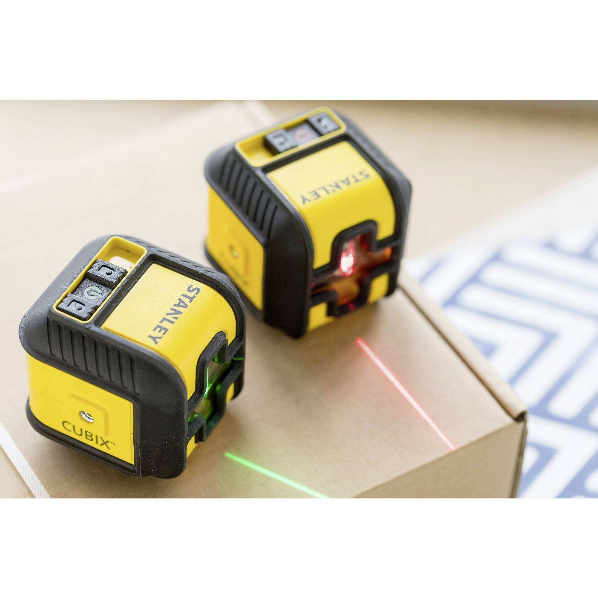 Zwei gelbe Laser-Wasserwaagen projizieren vertikale und horizontale grüne bzw. rote Laserlinien.  Sie befinden sich auf einem Karton.