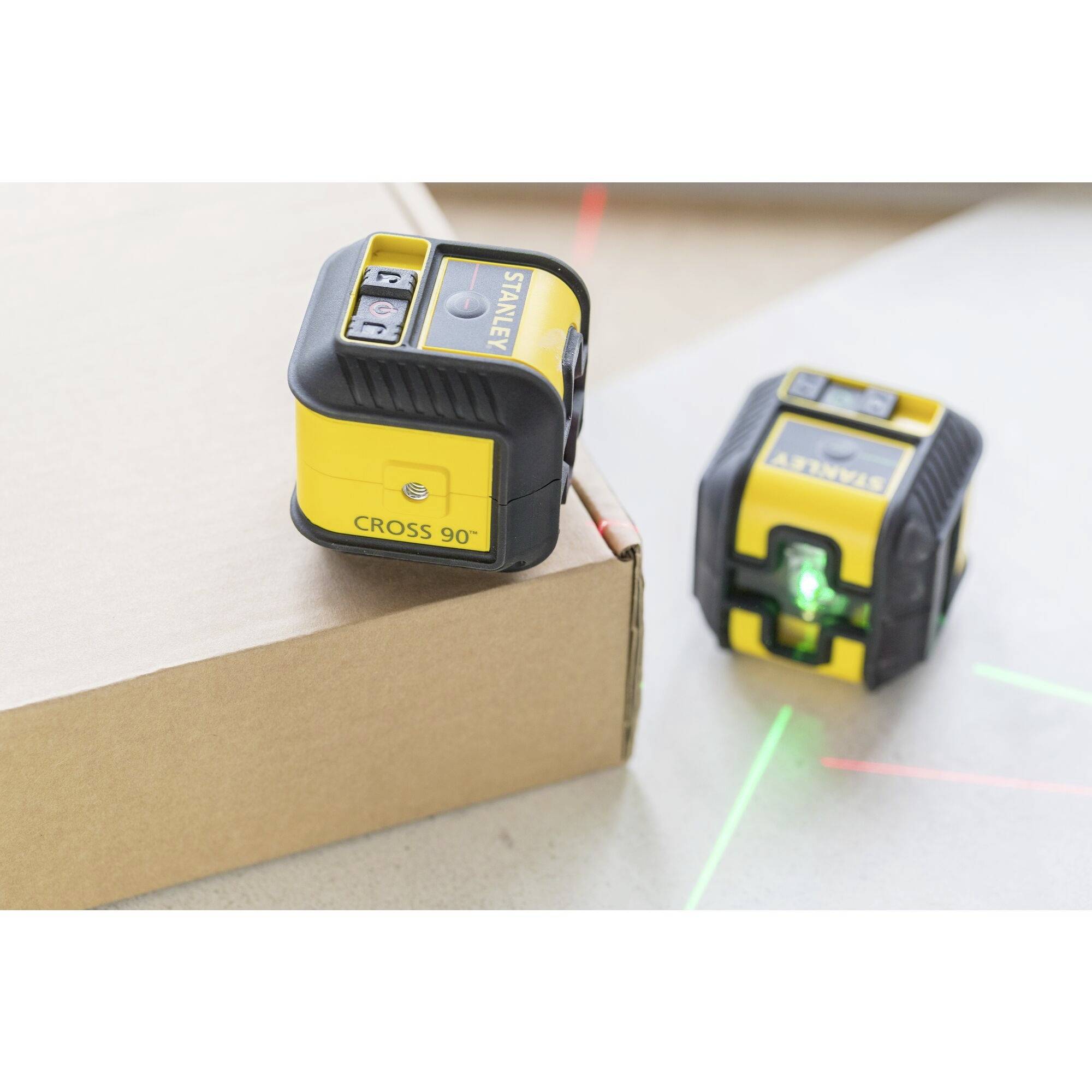 Zwei gelbe Lasernivelliergeräte auf einem Karton. Ein Gerät projiziert grüne und rote Laserlinien, während das andere ausgeschaltet ist.