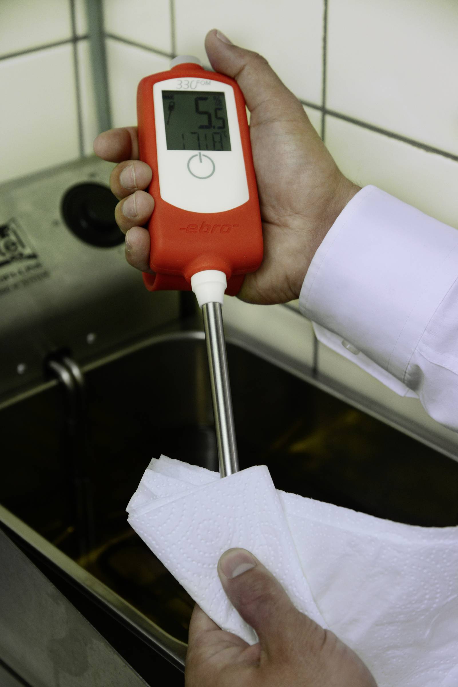 Person hält ein digitales Thermometer, das 56°C anzeigt, neben einem Becken mit Wasser; die Metallsonde wird sauber gewischt.