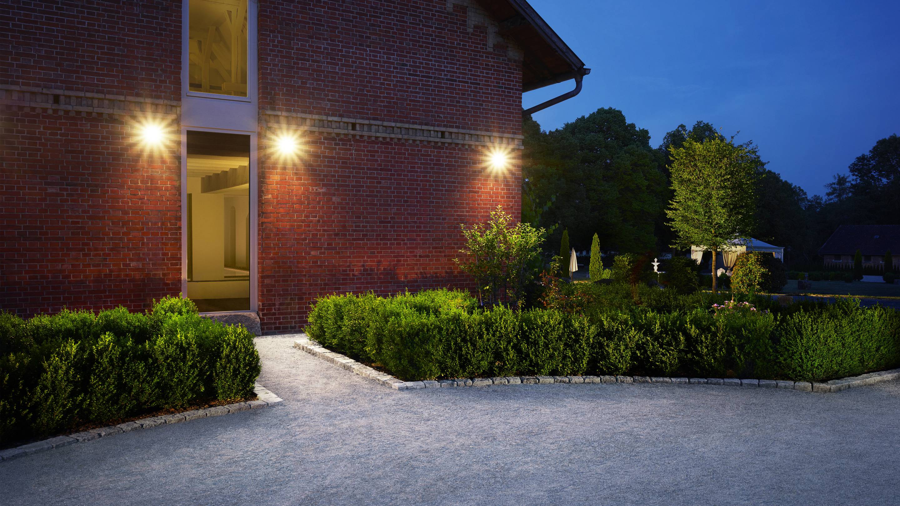 Ein beleuchteter Weg führt zu einem Backsteingebäude bei Abenddämmerung, umgeben von gepflegten Sträuchern und Bäumen im Hintergrund.