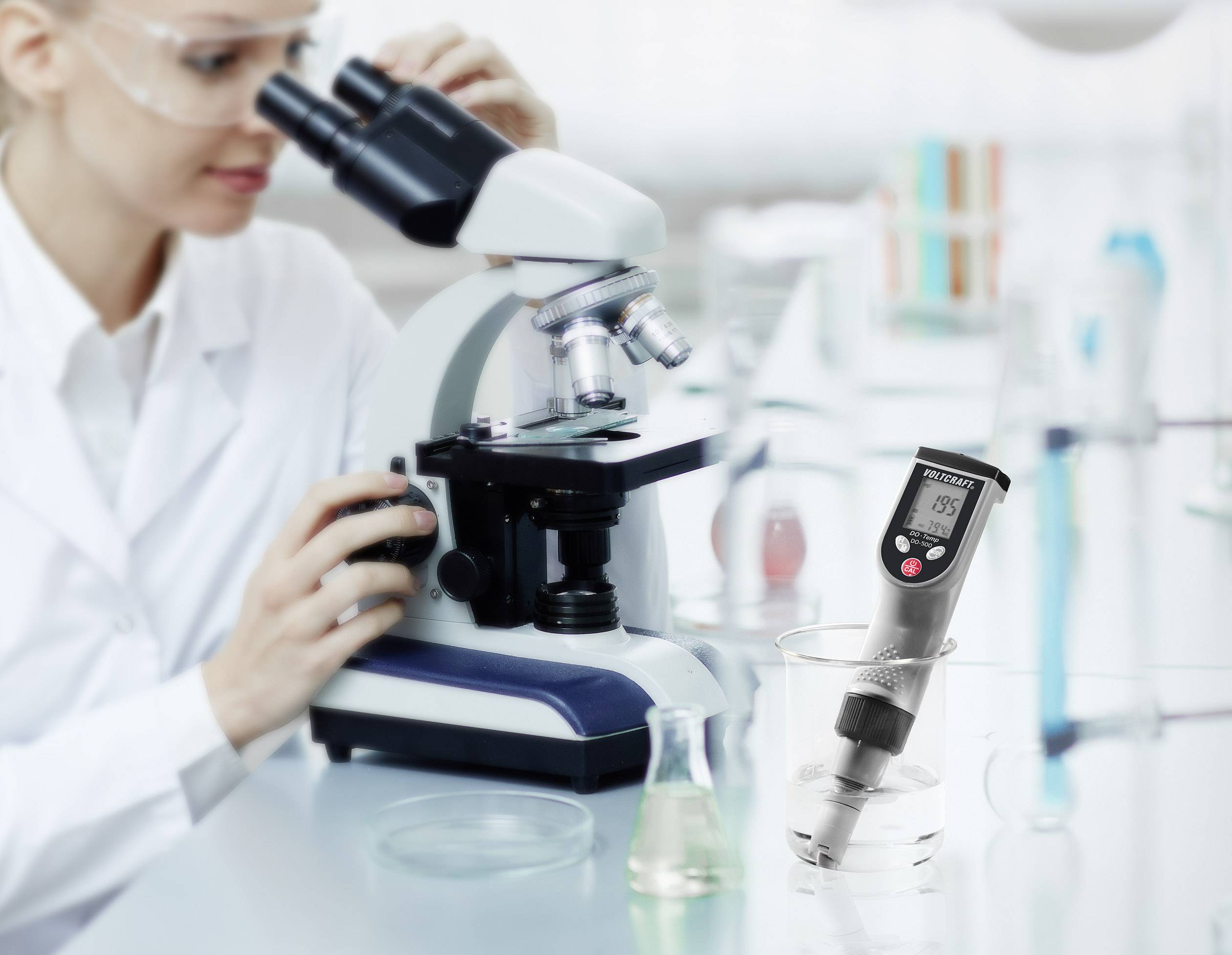 Eine Wissenschaftlerin betrachtet durch ein Mikroskop eine Probe. Ein digitales pH-Meter misst den pH-Wert einer Flüssigkeit daneben.