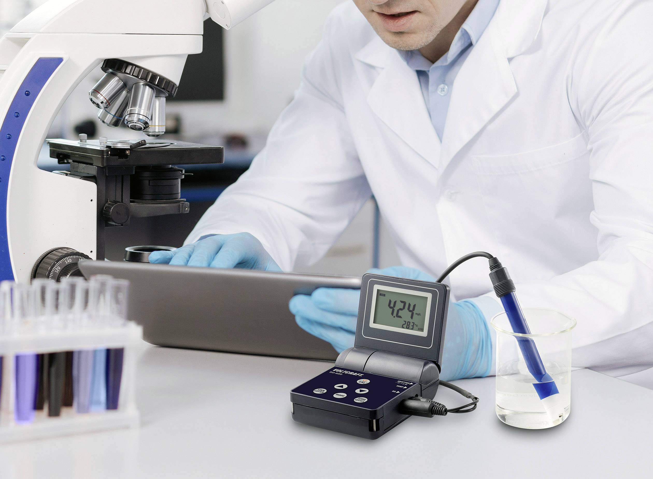 Ein Wissenschaftler im Labor arbeitet mit einem Mikroskop, daneben ein digitales Messgerät, das 42,4 anzeigt, und eine Elektrode in einem Becher.