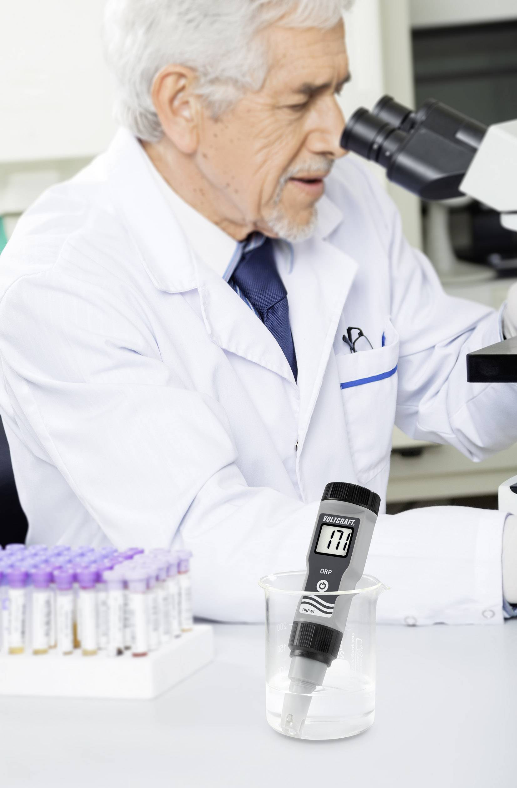 Ein Wissenschaftler untersucht Proben unter einem Mikroskop in einem Labor. Im Vordergrund steht ein Messgerät in einem Becherglas.