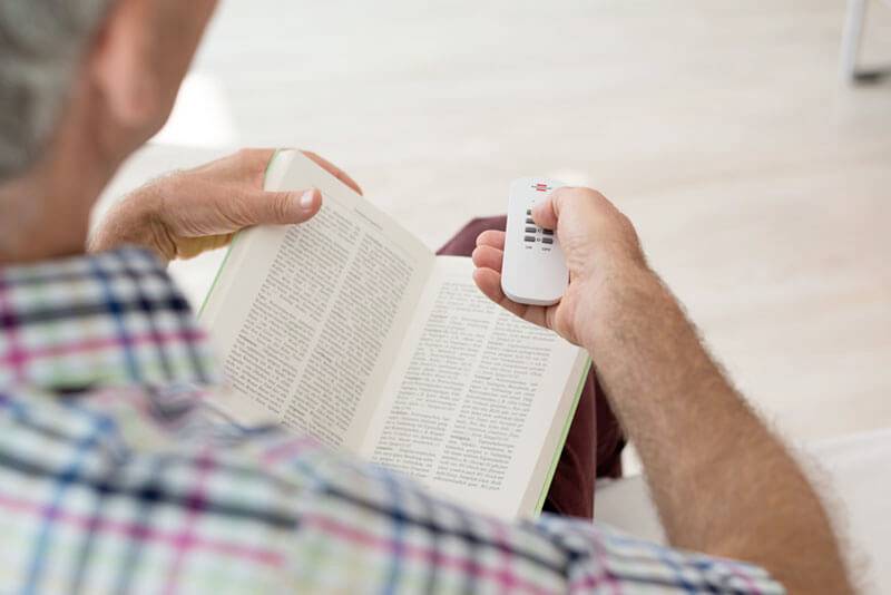 Eine Person sitzt in einem Raum und liest ein Buch, während sie eine Fernbedienung in der anderen Hand hält.
