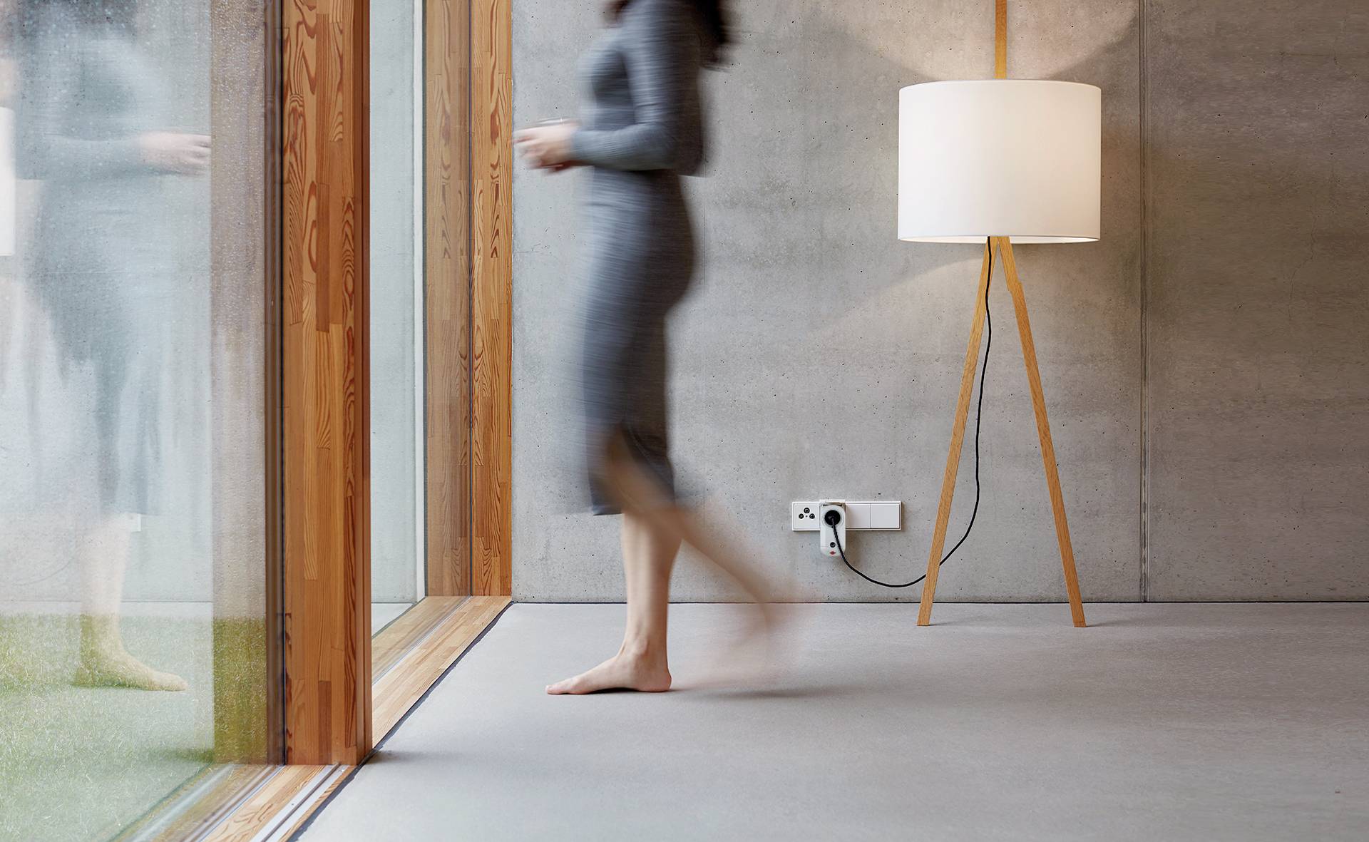 Eine Person geht barfuß in einem modernen Raum mit Betonwänden und einer Stehlampe am Fenster vorbei.