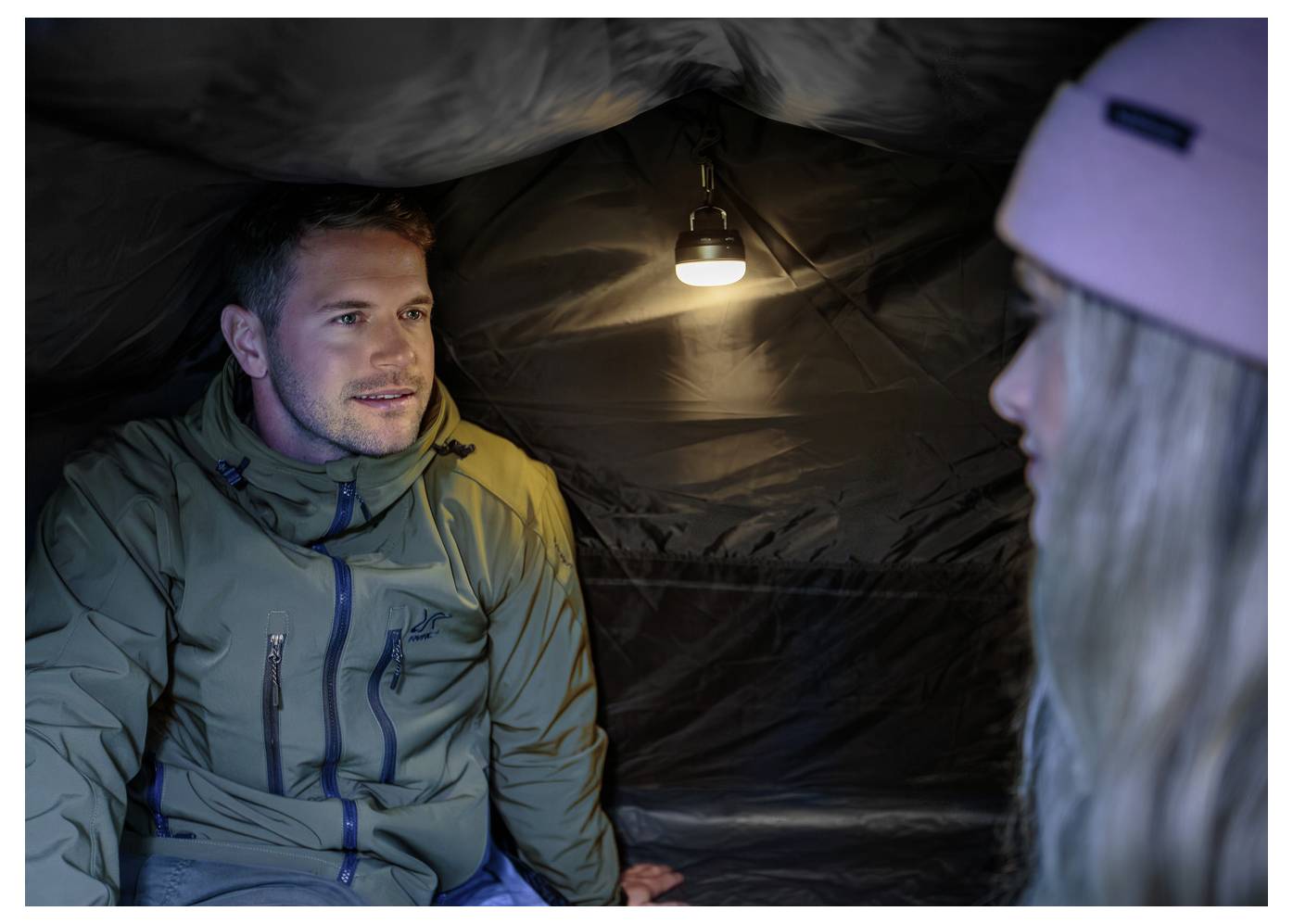 Zwei Menschen sitzen in einem kleinen Zelt, das von einer hängenden Lampe beleuchtet wird, und sprechen sich von Angesicht zu Angesicht gegenüber.