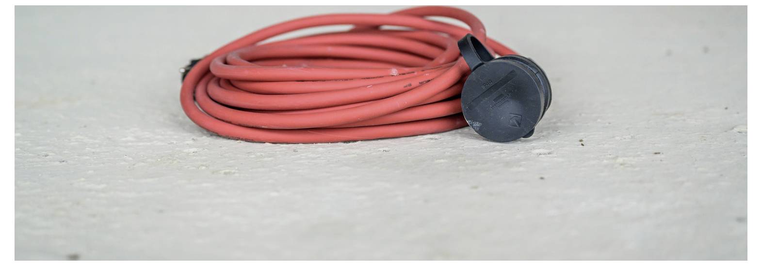 Aufgerolltes rotes Verlängerungskabel mit schwarzem Stecker, das auf einer hellgrauen Oberfläche ruht.
