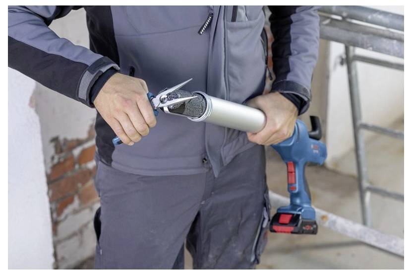 Eine Person schneidet die Düse einer Kartuschenpistole mit einem Cuttermesser zu und bereitet sie zur Verwendung vor. Sie steht in einem teilweise renovierten Raum.