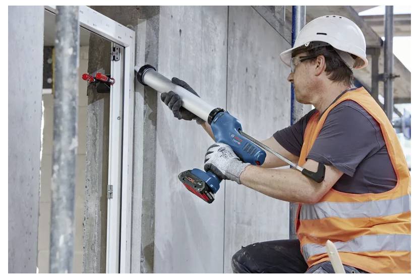 Ein Bauarbeiter mit Schutzhelm und orangefarbener Warnweste verwendet eine Kartuschenpistole, um einen Fensterrahmen auf einer Baustelle abzudichten.