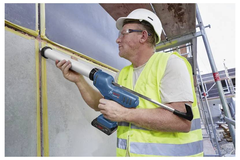 Ein Bauarbeiter mit Schutzhelm und Warnweste verwendet eine elektrische Kartuschenpistole, um Fugen in Wandpaneelen auf einer Baustelle zu verfugen.