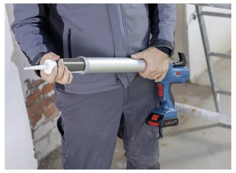 Person holding a cordless caulking gun, wearing work gear, standing at a construction site with exposed brick and ladder nearby.