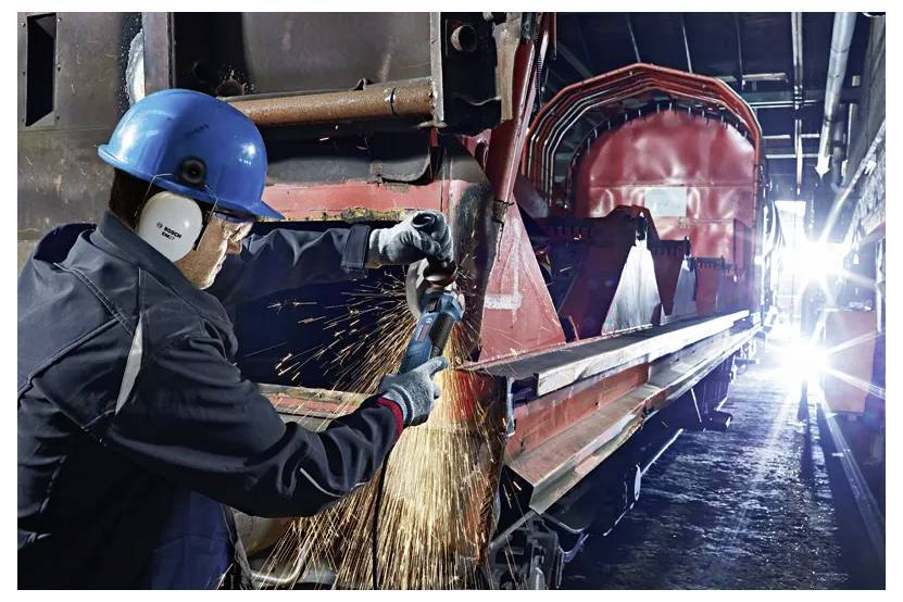 Ein Arbeiter in Schutzausrüstung schleift Metall in einem industriellen Umfeld, wobei helle Funken sprühen. Die Umgebung wirkt groß und mit Maschinen gefüllt.