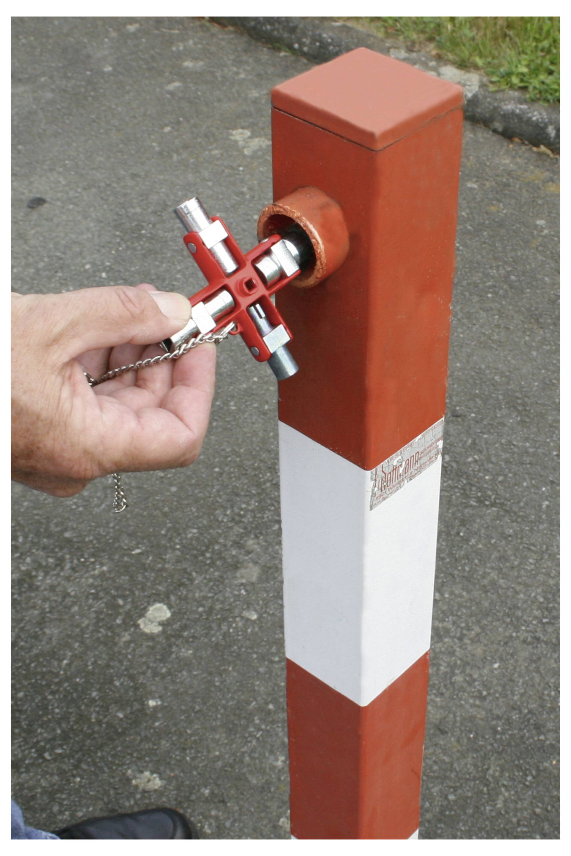 Eine Hand benutzt einen kreuzförmigen Schlüssel, um einen rot-weißen Poller auf einer gepflasterten Oberfläche zu entriegeln.