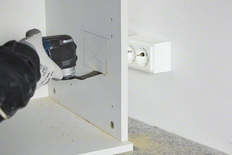 Eine Person schneidet mit einem Elektrowerkzeug ein rechteckiges Loch in ein weißes Holzregal nahe einer Wandsteckdose.