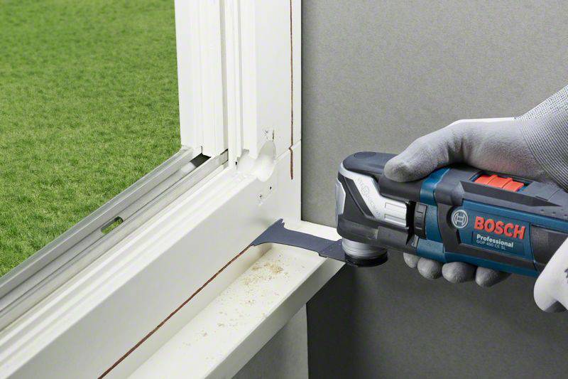 Eine Hand hält ein Bosch-Multifunktionswerkzeug und schneidet präzise in eine Fensterleiste. Draußen ist grüner Rasen sichtbar.