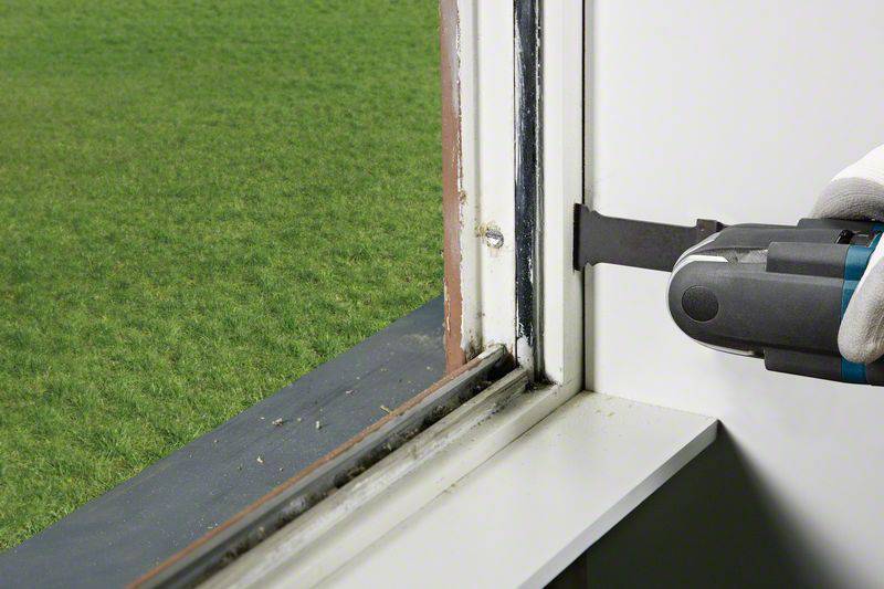Eine Person in weißen Handschuhen entfernt mit einem Werkzeug eine alte Fensterdichtung. Im Hintergrund ist Rasen sichtbar.