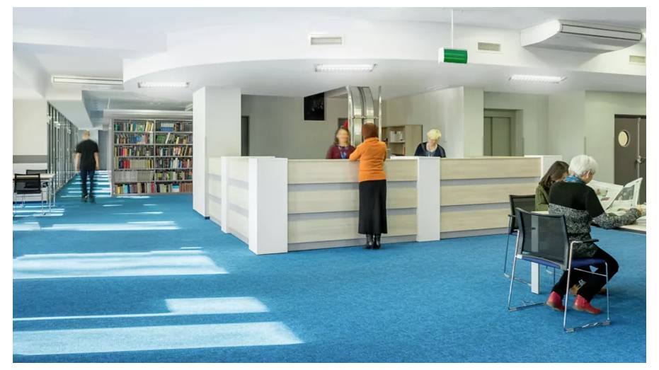 Ein Bibliotheksinterieur mit einer Ausleihtheke, die von zwei Personen besetzt ist. Eine Person steht an der Theke, während andere Bücher durchstöbern und lesen.