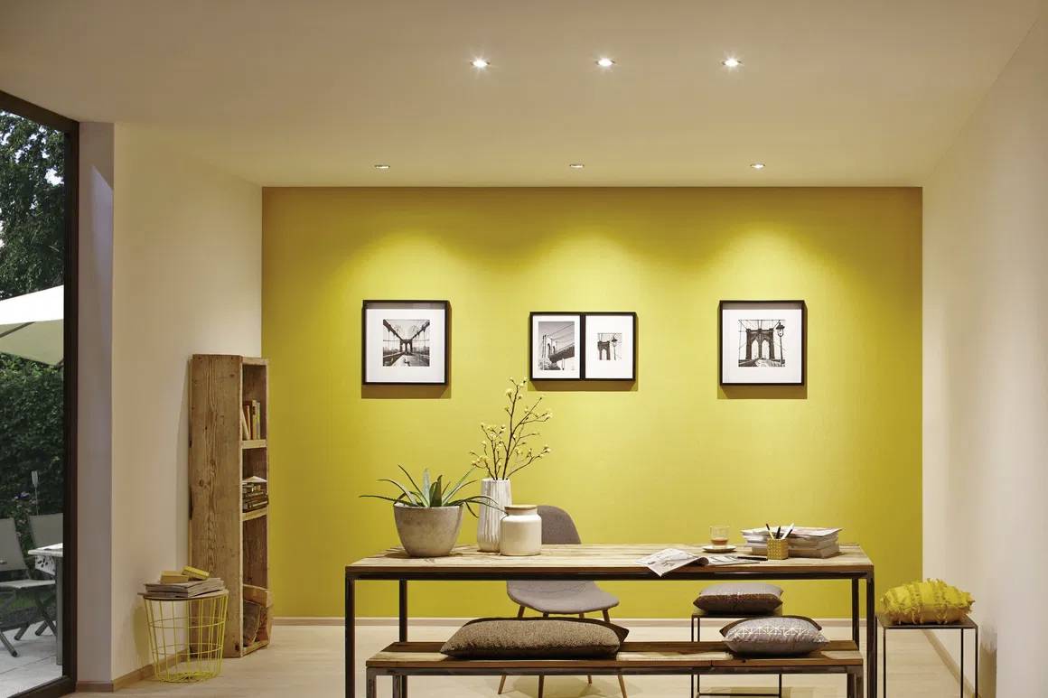 Ein helles Wohnzimmer mit gelber Akzentwand. Ein Holztisch ist in der Mitte platziert, dekoriert mit Pflanzen und Büchern. Drei Bilder hängen an der Wand.