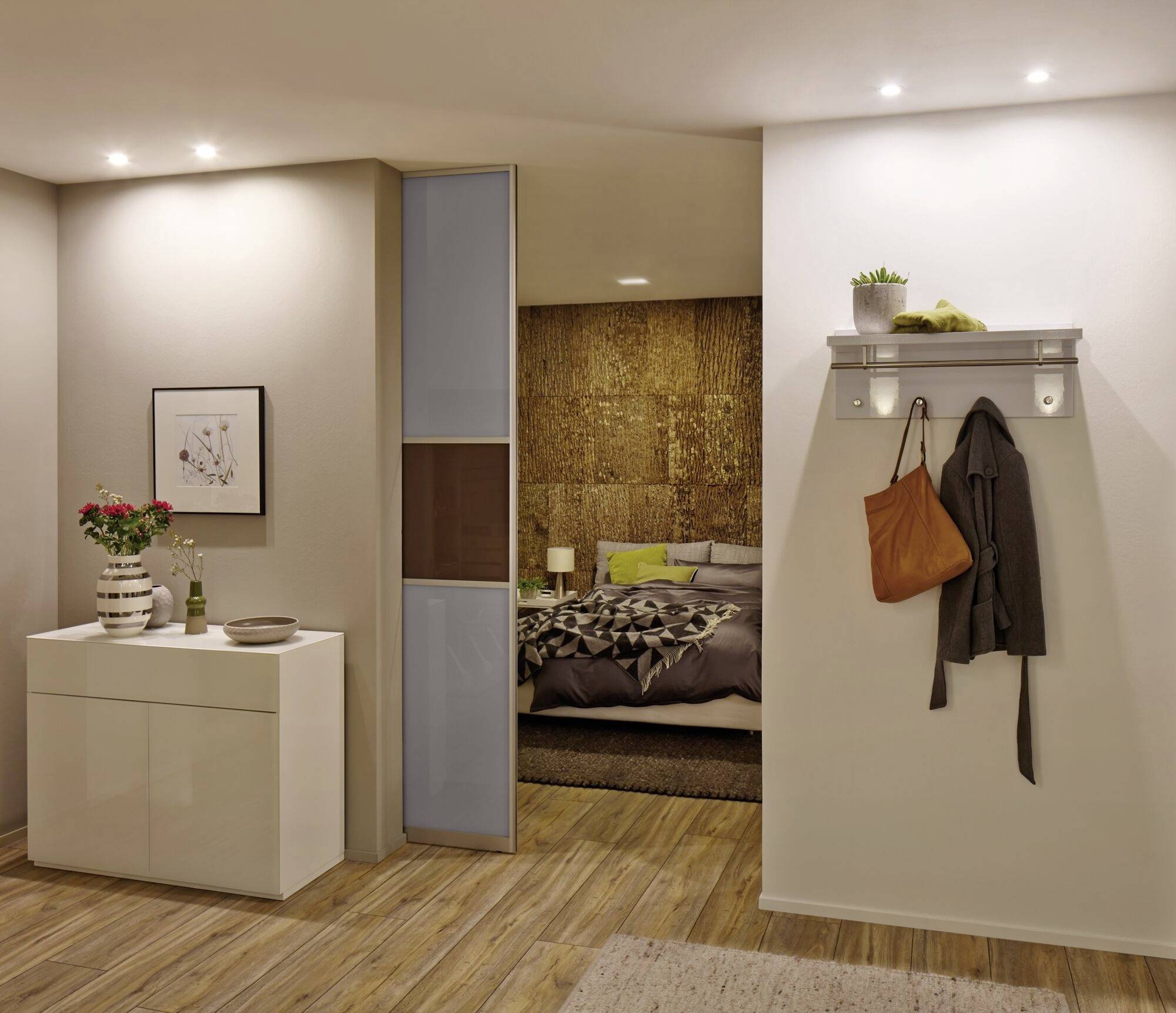 Ein modernes Schlafzimmer mit Holzfußboden. Links eine weiße Kommode mit Dekoration, rechts eine Tür mit Garderobe und Taschen.