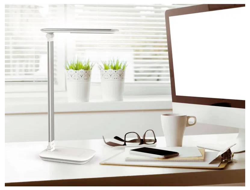 Eine moderne Schreibtischeinrichtung mit einer eleganten Lampe, einem Computermonitor, einer weißen Kaffeetasse, einer Brille und einem Notizblock auf einem hellfarbigen Tisch.