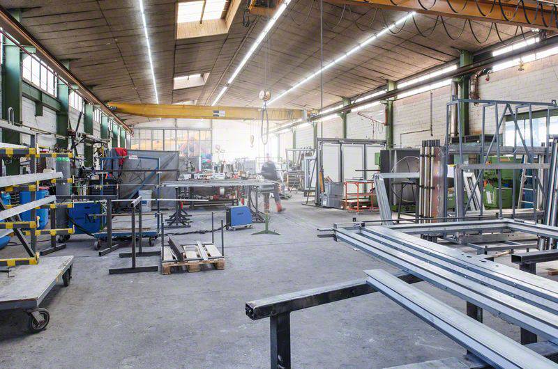 Werkstatt mit Metallschienen, Maschinen und Werkzeugen. Eine Person arbeitet im Hintergrund. Weitläufiger Raum mit hohen Decken.