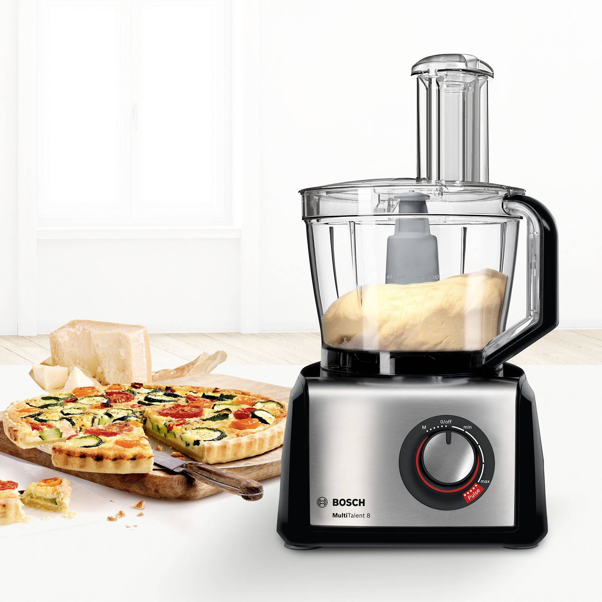 Küchenmaschine auf der Arbeitsplatte neben geschnittenem Brot und Pizza. Markiert Bosch, Modell MUM5, mit integriertem Mixer.