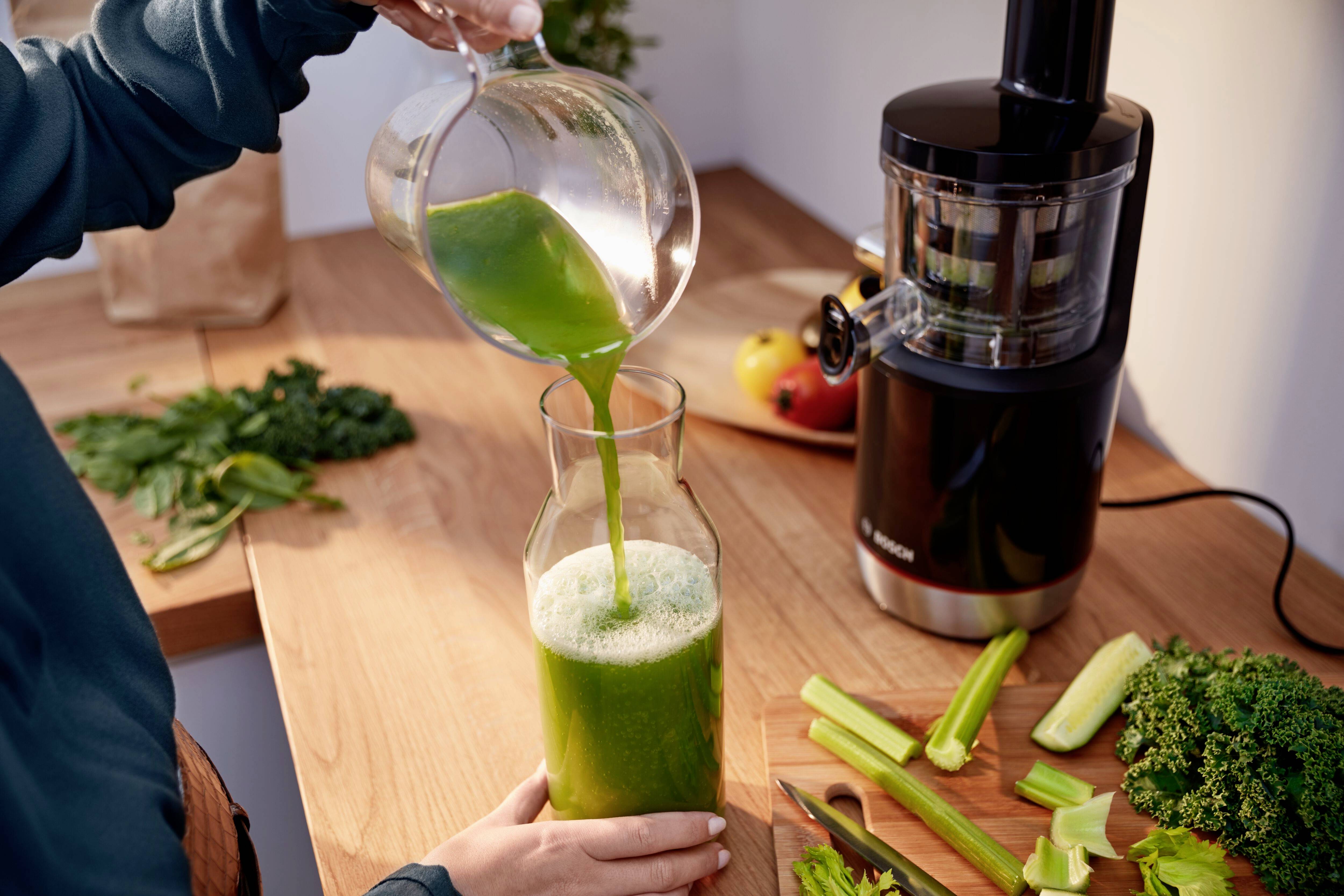 Person gießt grünen Smoothie in eine Glaskaraffe auf einer Holzarbeitsplatte. Im Hintergrund sind Obst, Gemüse und ein Entsafter zu sehen.