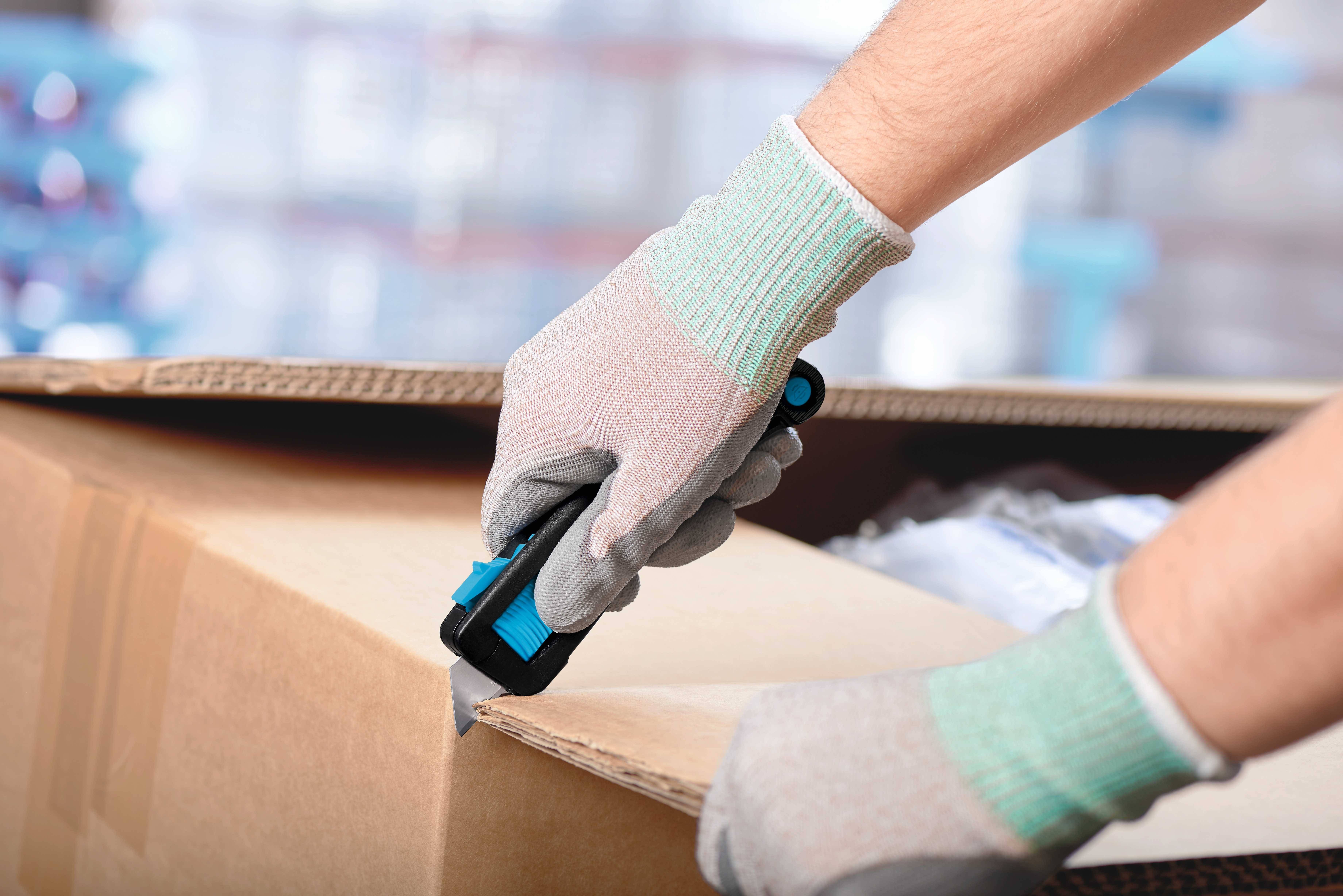 Eine Person mit grauen und grünen Handschuhen öffnet vorsichtig einen Karton mit einem blauen Cuttermesser.