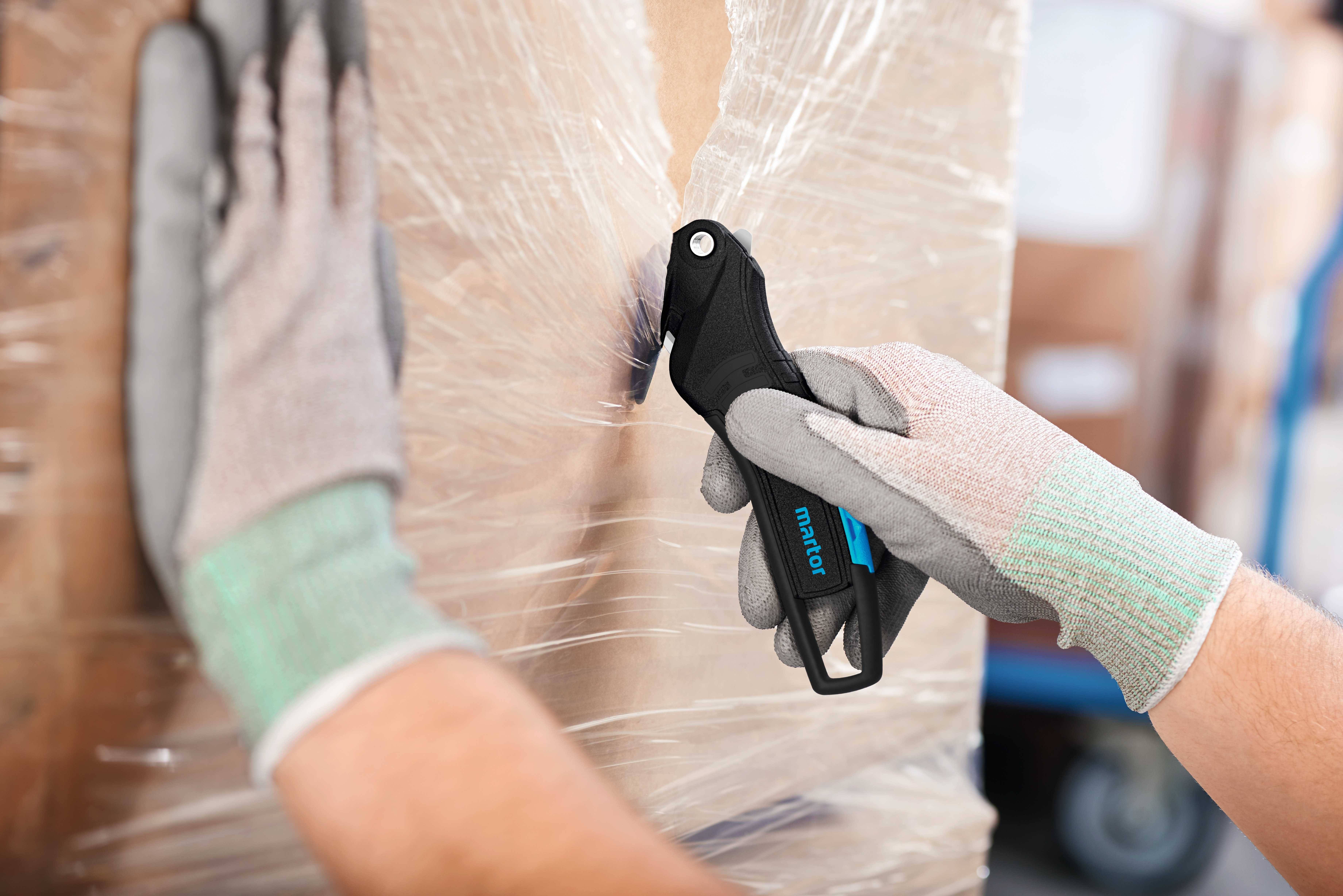 Eine Person schneidet mit einem Sicherheitsmesser Verpackungsfolie von einem Karton. Die Person trägt Arbeitshandschuhe.