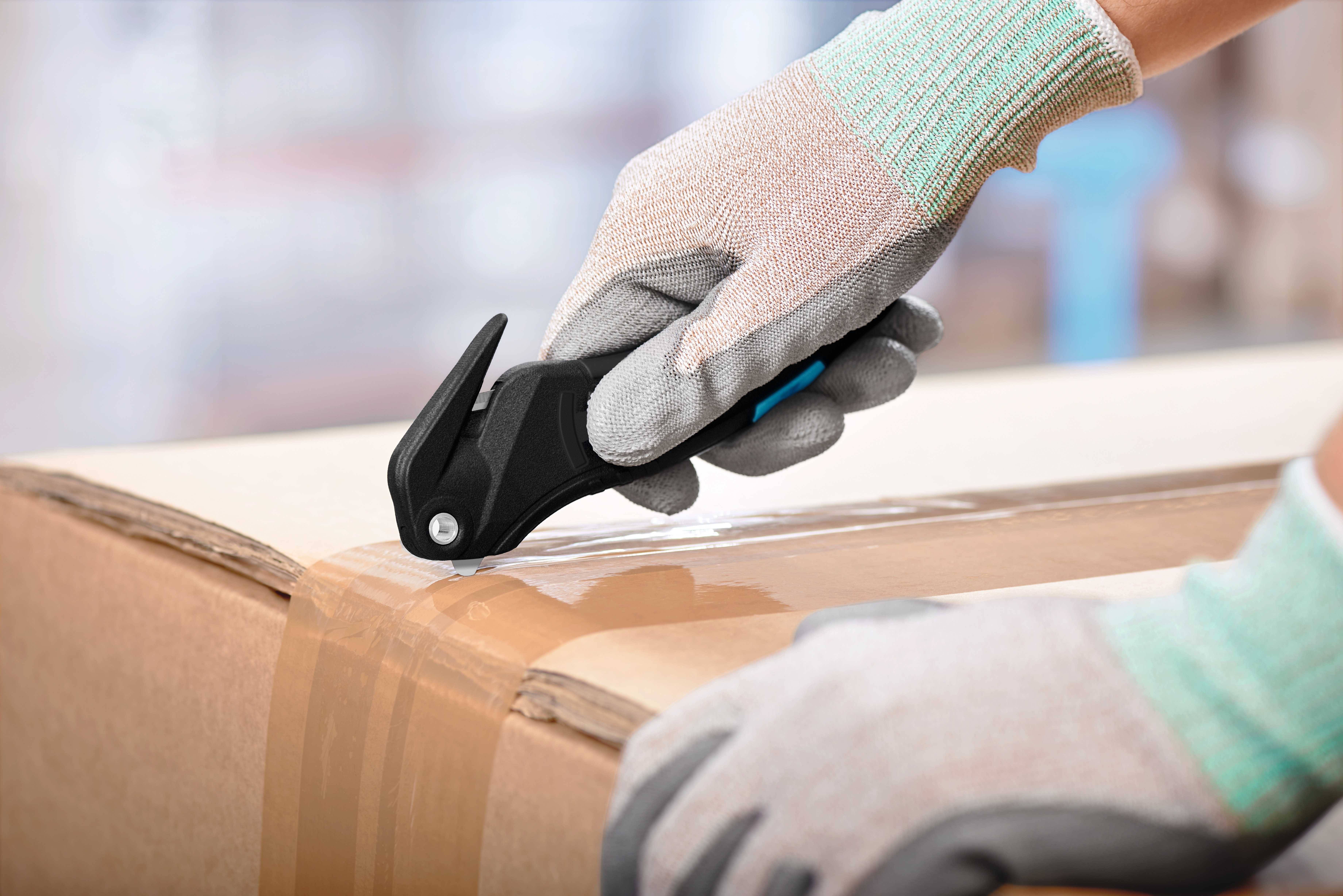 Eine Person mit Arbeitshandschuhen benutzt ein Sicherheitsmesser, um Klebeband auf einem Karton zu schneiden.