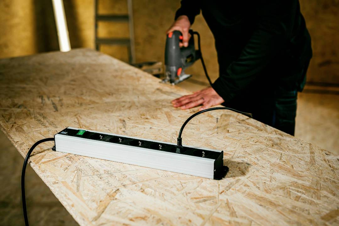 Person, die eine Stichsäge zum Schneiden von Sperrholz auf einer Werkbank in einer Werkstatt verwendet, mit einer Mehrfach-Steckdosenleiste im Vordergrund.