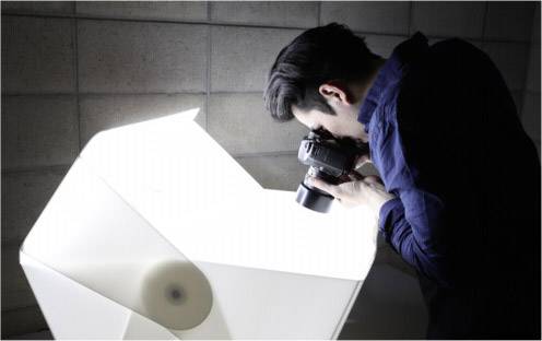 Eine Person fotografiert ein architektonisches Modell mit einer Kamera. Die Szene ist hell beleuchtet, die Umgebung wirkt professionell.