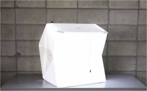 Ein hell beleuchteter Fotowürfel auf einem Tisch vor einer grauen Ziegelwand, genutzt für Produktfotografie.