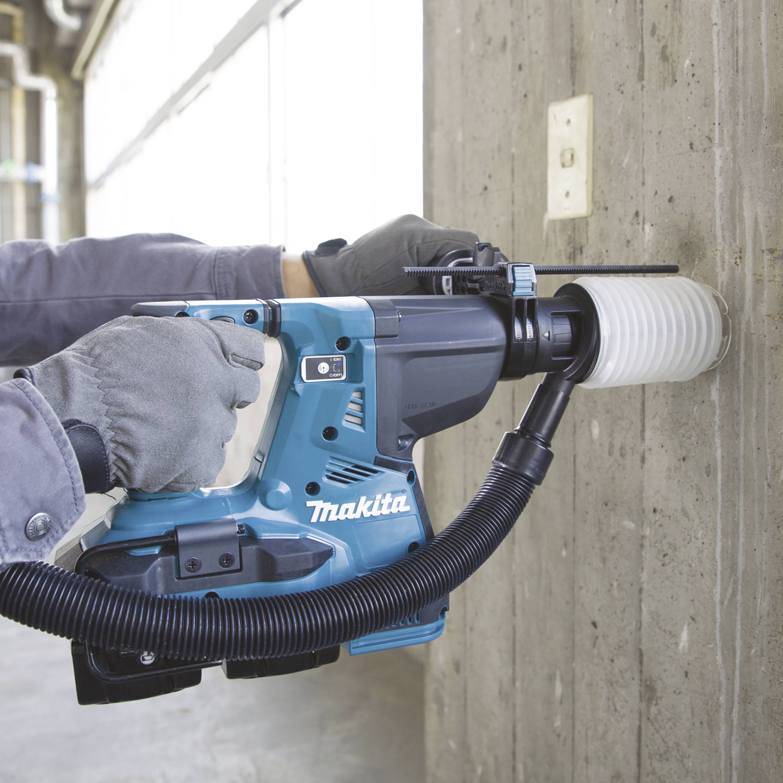 Eine Person nutzt einen blauen Bohrhammer, um ein Loch in eine Betonwand zu bohren. Der Bohrer hat einen Staubabsaugadapter.