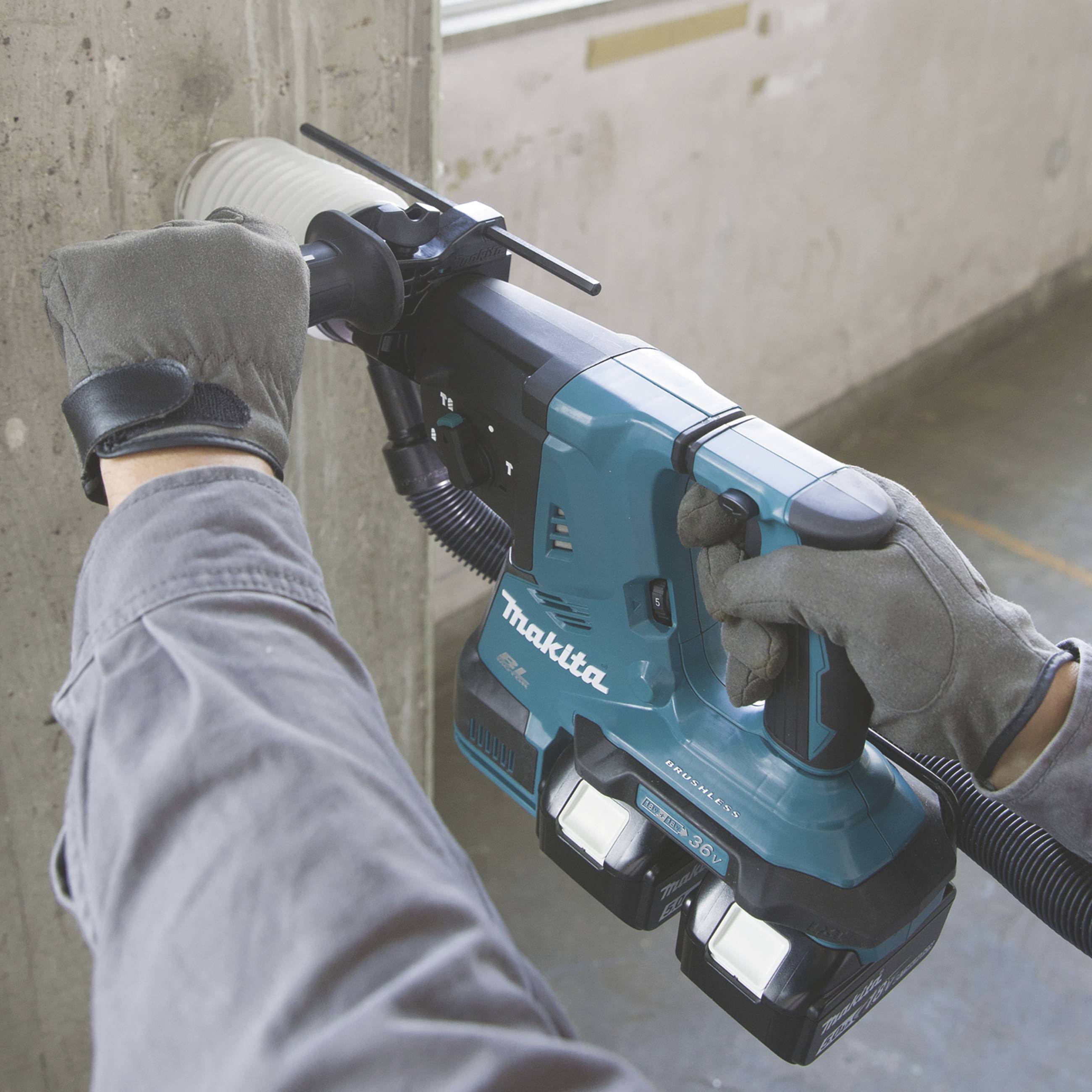 Ein Arbeiter bohrt mit einem blauen Bohrhammer in eine Betonwand. Der Bohrhammer hat zwei aufgesetzte Akkus und Staubabsaugung.