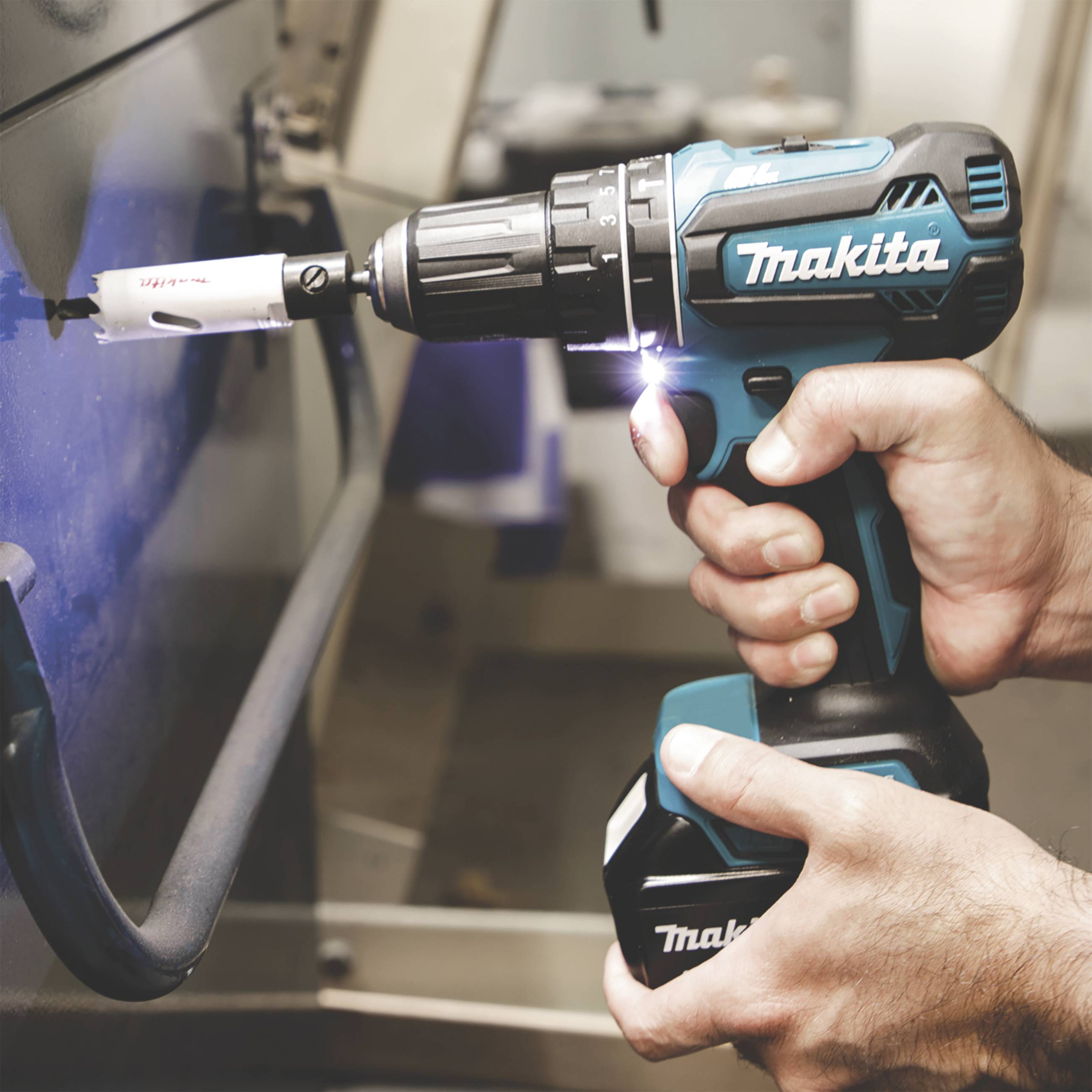 Person hält eine blaue Makita-Bohrmaschine mit einem Lochsägeaufsatz, während sie ein Metallstück bearbeitet.