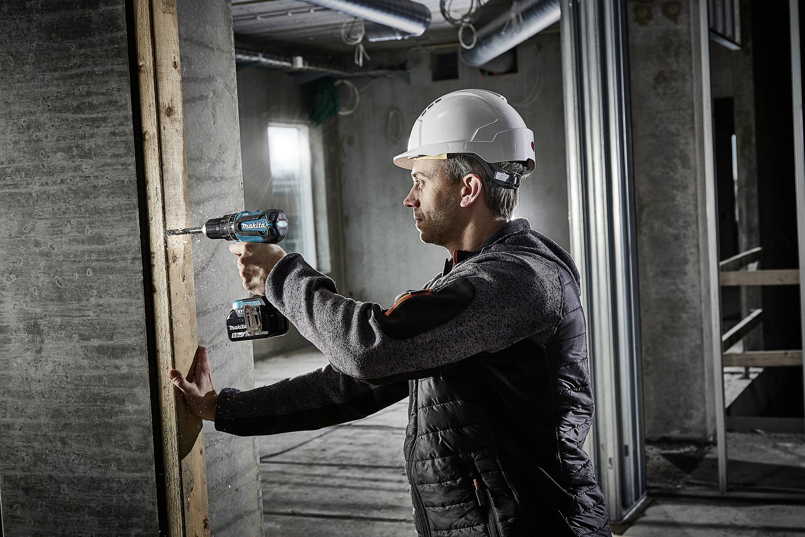 Ein Bauarbeiter mit Helm und Arbeitskleidung benutzt einen Akkuschrauber, um eine Schraube in eine Holzwand zu bohren, in einem Rohbau.