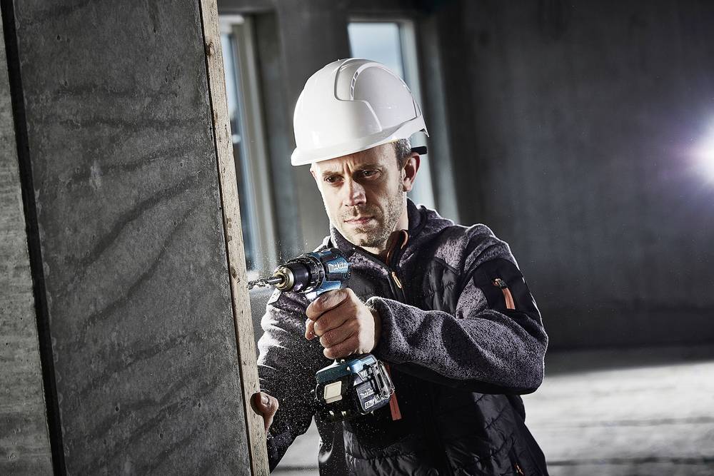 Ein Mann mit Schutzhelm verwendet einen Akkuschrauber, um ein Loch in eine Holzwand zu bohren. Hintergrund ist eine Baustelle.