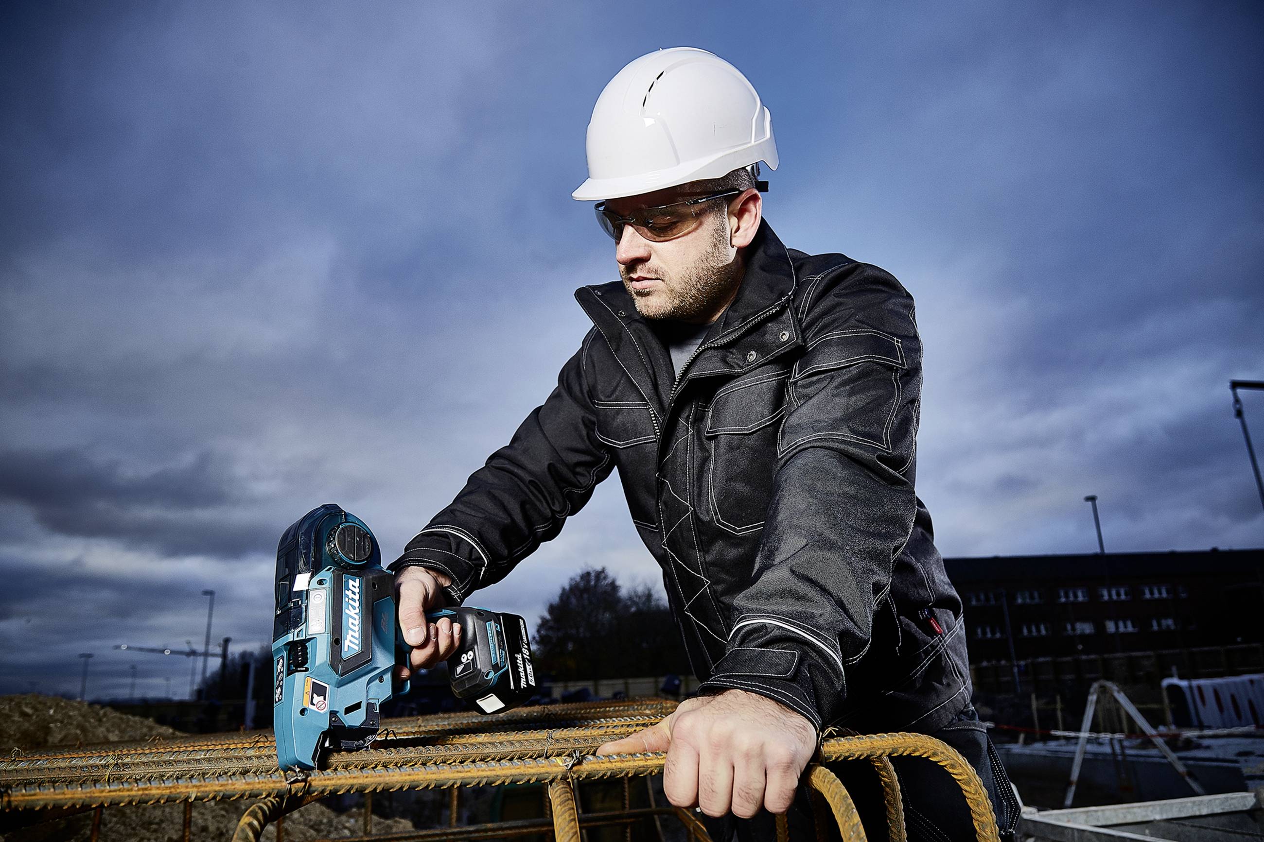 Ein Mann mit Schutzhelm und -brille arbeitet auf einer Baustelle, hält ein elektrisches Werkzeug und beugt sich über Stahlstangen.